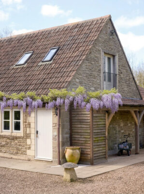 A corner view of a holiday cottage with flowering wisteria above the windows and doors at Great Ashley Farm in Bradford-On-Avon